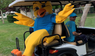 Maskot forklædt som gul fugl i blå trøje sidder i en golfvogn ved Camping Paradis Aubeterre i Frankrig.