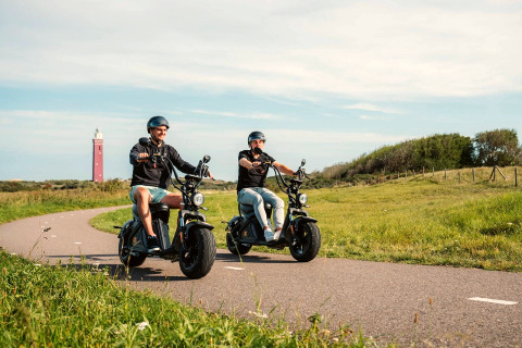 Deux personnes roulent en scooters électriques sur un chemin rural avec un phare rouge en arrière-plan.