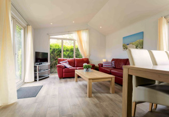 Helles Wohnzimmer in einer Hütte mit roten Sofas, Holztisch, Fernseher und großen Fenstern zum Garten.