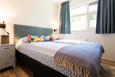 Dormitorio con cama doble, cojines coloridos, manta azul y grandes ventanas en alojamiento glamping.