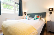 Luminoso dormitorio glamping con cama acogedora, cojines coloridos, grandes ventanas y luz moderna.
