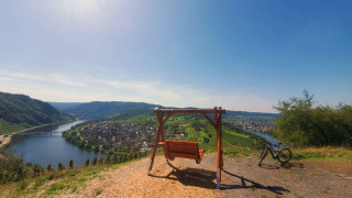 Scenic view near Kröv, Germany, showing a hilltop swing, bicycle, and the Moselle River in sunshine.