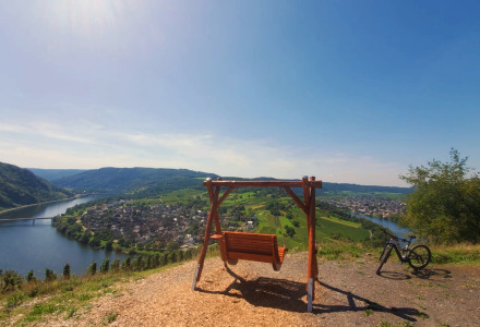 Scenic view near Kröv, Germany, showing a hilltop swing, bicycle, and the Moselle River in sunshine.