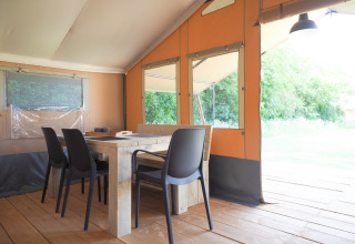 Binnenkant van een safaritent met houten tafel, zwarte stoelen en grote ramen met zicht op de natuur.