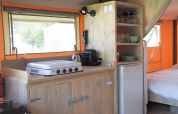 Kitchen inside a safari tent featuring a gas stove, coffee maker, fridge, and shelves with tableware.