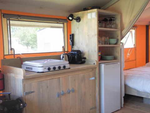 Cocina en una tienda safari con estufa de gas, cafetera, nevera y estantes con vajilla.
