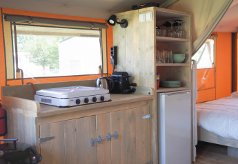 Kitchen inside a safari tent featuring a gas stove, coffee maker, fridge, and shelves with tableware.