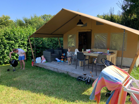 Safari telt med terrasse, børn leger og en voksen griller udenfor på solrig dag omgivet af grønt.