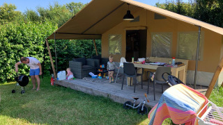 Tienda de safari con terraza, niños jugando y adulto asando afuera en un día soleado rodeado de vegetación.