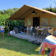 Safari telt med terrasse, børn leger og en voksen griller udenfor på solrig dag omgivet af grønt.