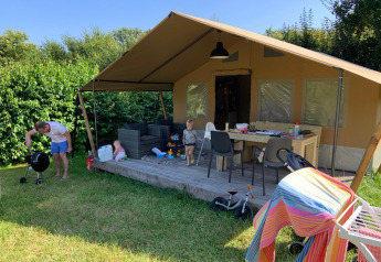 Tienda de safari con terraza, niños jugando y adulto asando afuera en un día soleado rodeado de vegetación.
