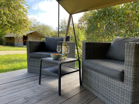 Cozy outdoor sitting area with wicker chairs and table, in front of a safari tent at Camping Paradis La Bretonniere, France.