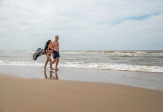 Par går barfodet langs stranden ved havet under ophold på feriepark med glamping i baggrunden.