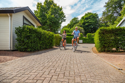 Twee mensen fietsen op een betegeld pad tussen huisjes en heggen in een glamping vakantiepark.
