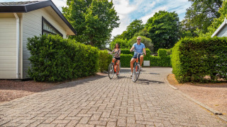 Dos personas pasean en bicicleta por un camino pavimentado entre cabañas y arbustos en un glamping.