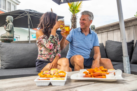 Deux personnes trinquent et dégustent des snacks en terrasse dans un parc de vacances avec glamping.