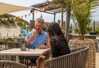 Koppel smult van ijsje op terras aan een vakantiepark, omringd door duinen en potplanten.