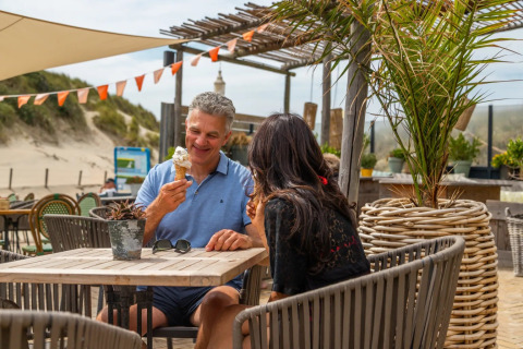 Et par nyder en is ved et bord i ferieparkens udendørs café med sanddyner og planter i baggrunden.
