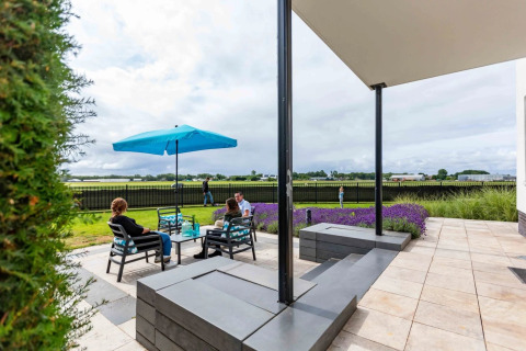 Personas relajándose bajo una sombrilla azul en una terraza con vistas a campos en un parque de glamping.