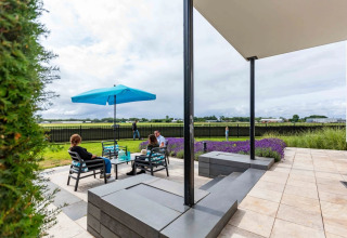 Personer slapper af under en blå parasol på en terrasse med udsigt til marker i en feriepark med glamping.