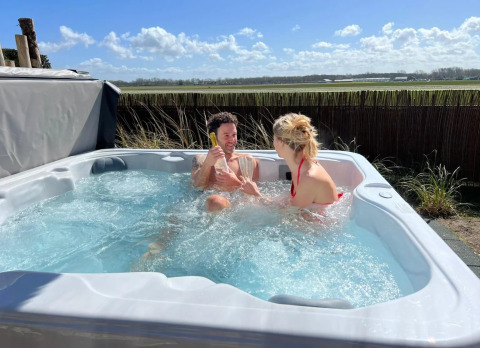 Twee mensen ontspannen in een bubbelbad buiten op een glamping, met uitzicht op groene velden en zon.
