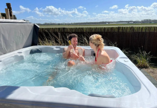 Twee personen genieten van een buitenjacuzzi bij een glamping, met zicht op groene weiden en heldere lucht.