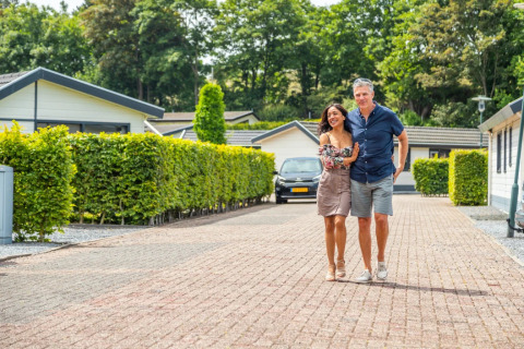 Una pareja camina abrazada por un sendero de adoquines entre cabañas modernas y setos en un parque vacacional.
