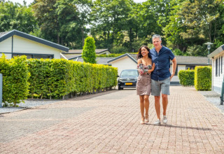 Una pareja camina abrazada por un sendero de adoquines entre cabañas modernas y setos en un parque vacacional.