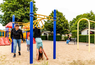 Børn leger på legepladsen i en feriepark med glamping, omgivet af legeudstyr og grønne træer.