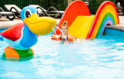 Lille pige leger i swimmingpool med regnbuerutsjebane og stor and ved et feriecenter med glamping.
