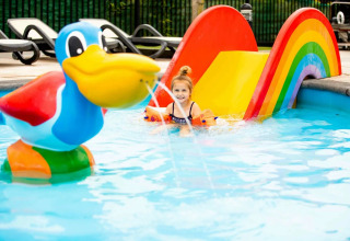 Niña pequeña jugando en piscina con tobogán arcoíris y pato grande en parque vacacional con glamping.