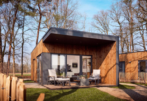 Cabaña moderna de madera con terraza y sillones, rodeada de árboles altos en un entorno tranquilo.