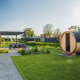 Moderne lodge med træsauna i haven og siddeområde udendørs under pergola omgivet af grønne områder.