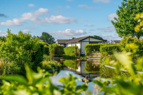 Cabaña de glamping junto a un canal, rodeada de vegetación y jardines en un parque vacacional soleado.