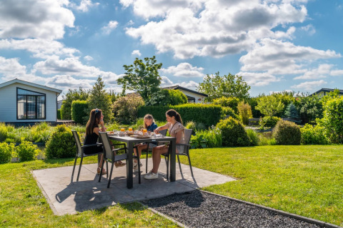 Three people enjoy breakfast outdoors at a table in a holiday park with glamping, surrounded by greenery.