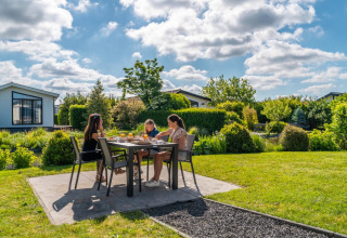 Drie personen ontbijten buiten aan een tafel in een vakantiepark met glamping, omringd door groen.