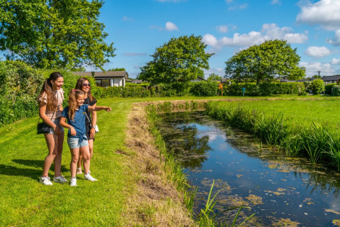 En familie nyder naturen ved en lille sø i en feriepark, perfekt til glamping og udendørs aktiviteter.