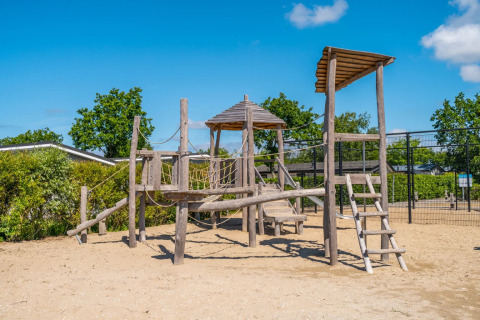 Holzspielplatz im Ferienpark auf sandigem Boden mit Bäumen, blauem Himmel und weißen Wolken.