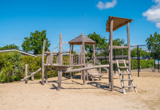 Houten speeltoestel op zand in een vakantiepark, omringd door bomen en een stralend blauwe lucht.
