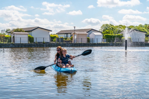 Madre e hijo remando en kayak en un lago junto a alojamientos glamping y una fuente en un parque vacacional.