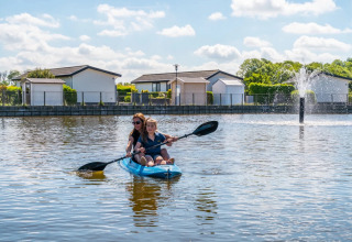 Madre e hijo remando en kayak en un lago junto a alojamientos glamping y una fuente en un parque vacacional.
