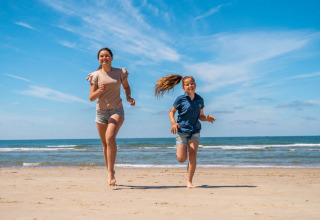 Two people running barefoot on a sandy beach at a holiday park offering glamping, enjoying sunshine.