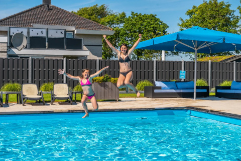 Two women jump into an outdoor pool at a holiday park offering glamping, sun loungers, and umbrellas.