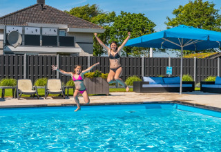 Twee vrouwen springen in een buitenzwembad bij een vakantiepark met glamping, ligbedden en blauwe parasols.