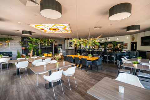 Modern restaurant in een vakantiepark met houten tafels, groene planten en centrale bar voor glamping.