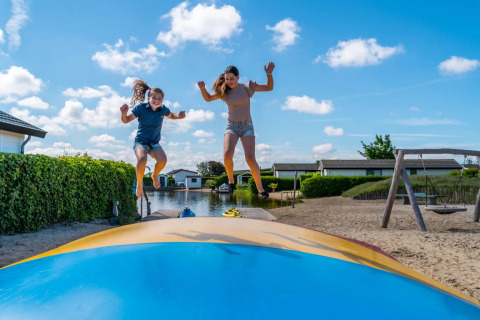 To børn hopper på en oppustelig pude ved en feriepark med hytter og sand på en solrig dag.