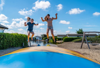 Two children jump on an inflatable pillow at a holiday park with cabins and sandy area on a sunny day.
