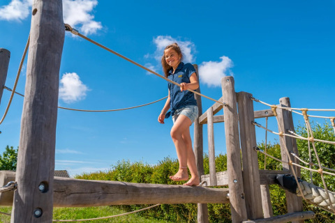 Et barn leger på en trælegeplads under en blå himmel i en feriepark med glamping faciliteter.