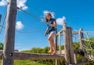 Een kind geniet op een houten speeltuig in een vakantiepark met glamping, onder een heldere lucht.
