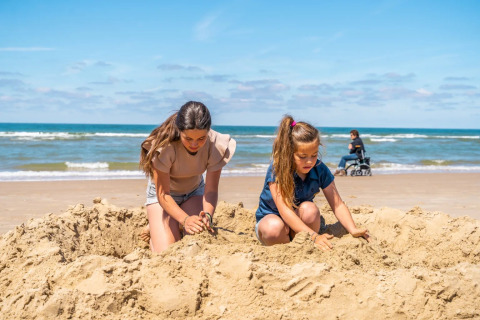 Twee meisjes bouwen in het zand aan het strand van een glampingpark, vrouw in rolstoel op de achtergrond.
